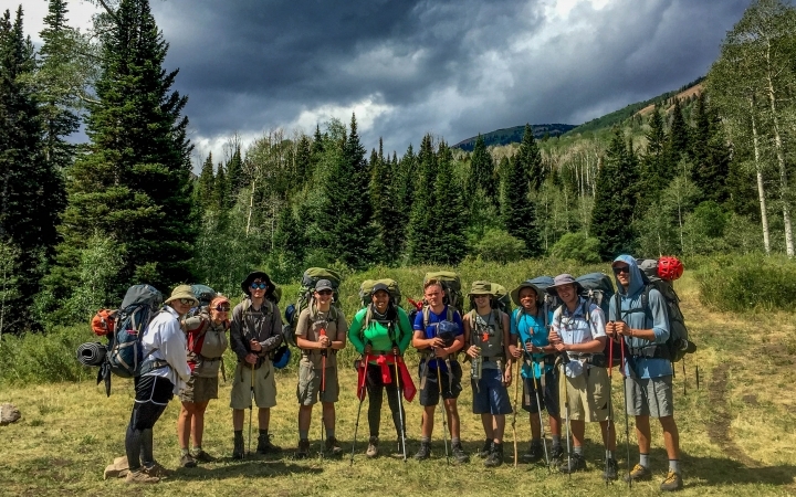 A group of gap year students wearing backpacks pose for a photo. There are evergreen trees in the background.